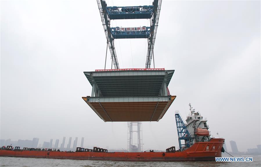 A 1,000-tonne steel beam is lifted to be installed at the construction site of the Yangsigang Bridge across the Yangtze River in Wuhan, capital of central China\'s Hubei Province, on Nov. 15, 2018. Construction of a double-deck suspension bridge with the longest span in the world is expected to be completed in October 2019, said an engineer working on the project. The bridge, with a 1,700-meter-long main span across the Yangtze River in Wuhan, saw the first steal beam installed for the bridge deck Thursday. (Xinhua/Cheng Min)