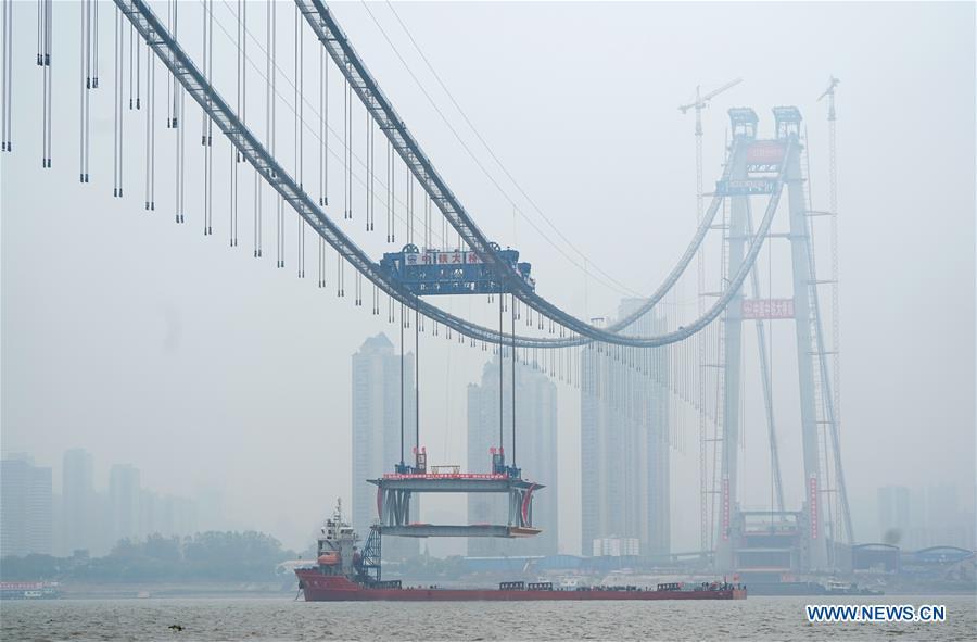 A 1,000-tonne steel beam is lifted to be installed at the construction site of the Yangsigang Bridge across the Yangtze River in Wuhan, capital of central China\'s Hubei Province, on Nov. 15, 2018. Construction of a double-deck suspension bridge with the longest span in the world is expected to be completed in October 2019, said an engineer working on the project. The bridge, with a 1,700-meter-long main span across the Yangtze River in Wuhan, saw the first steal beam installed for the bridge deck Thursday. (Xinhua/Cheng Min)