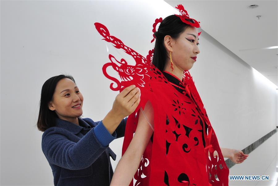 Craftswoman Liang Ying (L) helps a cheongsam enthusiast put on her papercutting work of cheongsam at her studio in Liaocheng, east China\'s Shandong Province, Jan. 30, 2019. Liang Ying made 18 papercutting works of cheongsam to greet the upcoming Spring Festival, which falls on Feb. 5 this year. (Xinhua/Xu Wenhao)