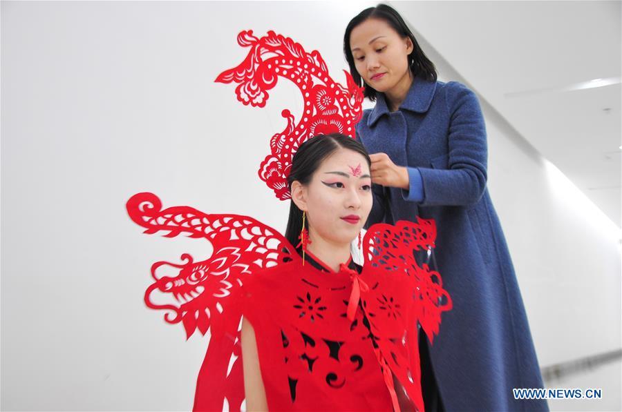 Craftswoman Liang Ying (R) helps a cheongsam enthusiast put on her papercutting work of cheongsam at her studio in Liaocheng, east China\'s Shandong Province, Jan. 30, 2019. Liang Ying made 18 papercutting works of cheongsam to greet the upcoming Spring Festival, which falls on Feb. 5 this year. (Xinhua/Xu Wenhao)