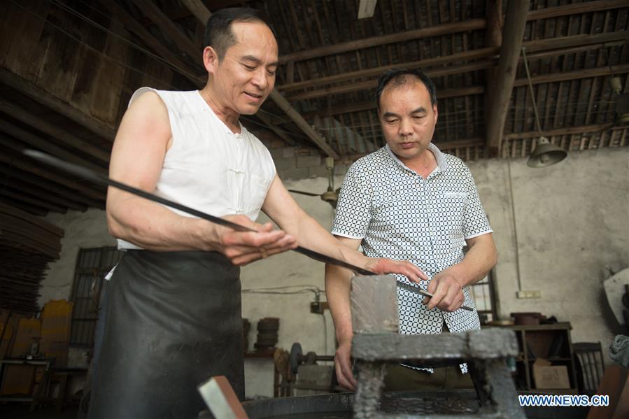 Zheng Guorong (L) and his co-worker Wu Changyou discuss sword burnishing skills at Guyue Sword Studio in Longquan, east China\'s Zhejiang Province, May 21, 2019. For centuries, Longquan has been famed for its sword making industry. Zheng Guorong, 55, an intangible cultural heritage inheritor in Longquan Sword making, has been making sword for more than 30 years. Zheng regards sword as a symbol of spirit and devotes all to his career with awe. In his studio, the whole process of making a single sword takes two to three months, with steps including forming an idea, designing the sketch, preparing material and the final making and assembling. After setting up his studio, Zheng has trained more than 20 apprentices, who have all become sword smiths of great reputation. The old sword smith insists that Longquan sword, with a history of more than 2,500 years, deserves to be carried on in our time, both materially and spiritually. (Xinhua/Weng Xinyang)